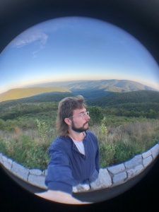 We see Andrew through a narrow lens standing on a stone wall and staring off to the side in front of a vast rolling landscape of mountains and hills. 